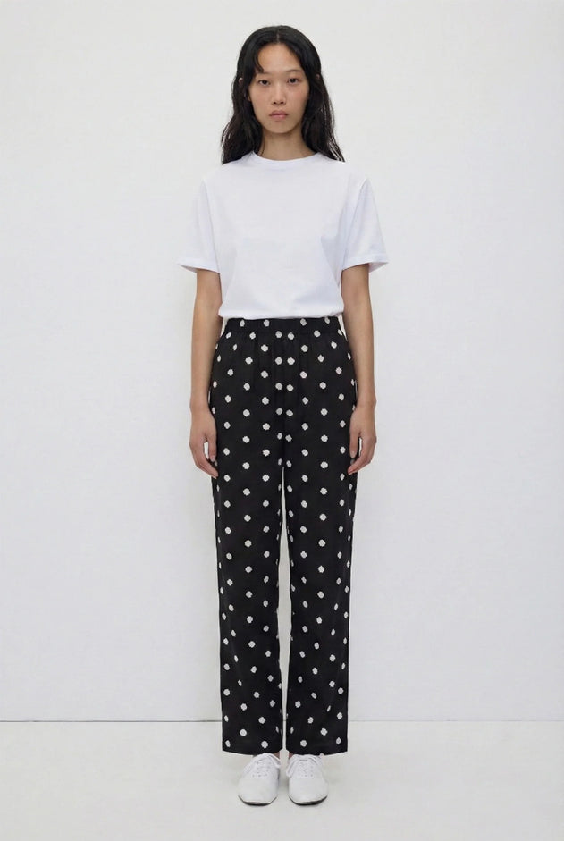 A model stands against a plain white studio background wearing a white short-sleeve crewneck t-shirt and black wide-leg trousers with a white polka-dot pattern, paired with white sneakers.
