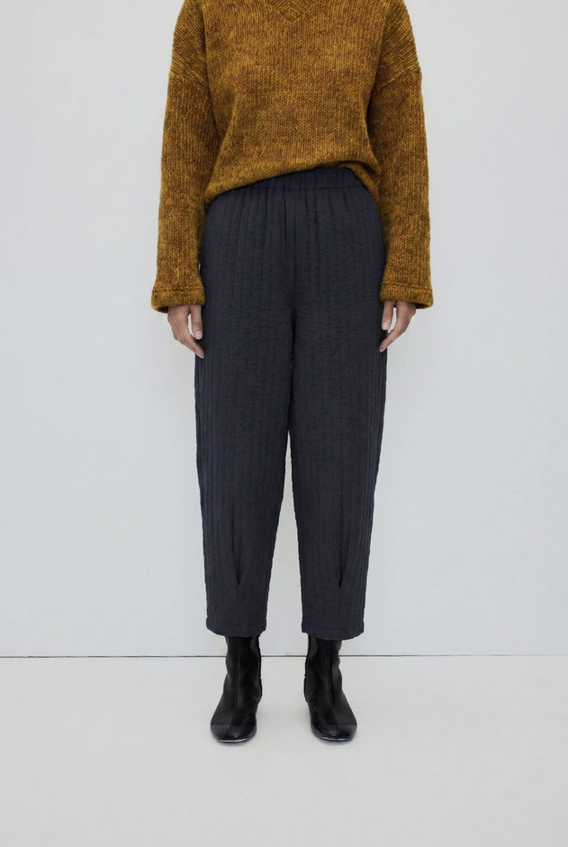 A model is shown from the chest down wearing dark charcoal gray pleated cropped trousers, a textured mustard yellow sweater, and black ankle boots against a plain white studio background.