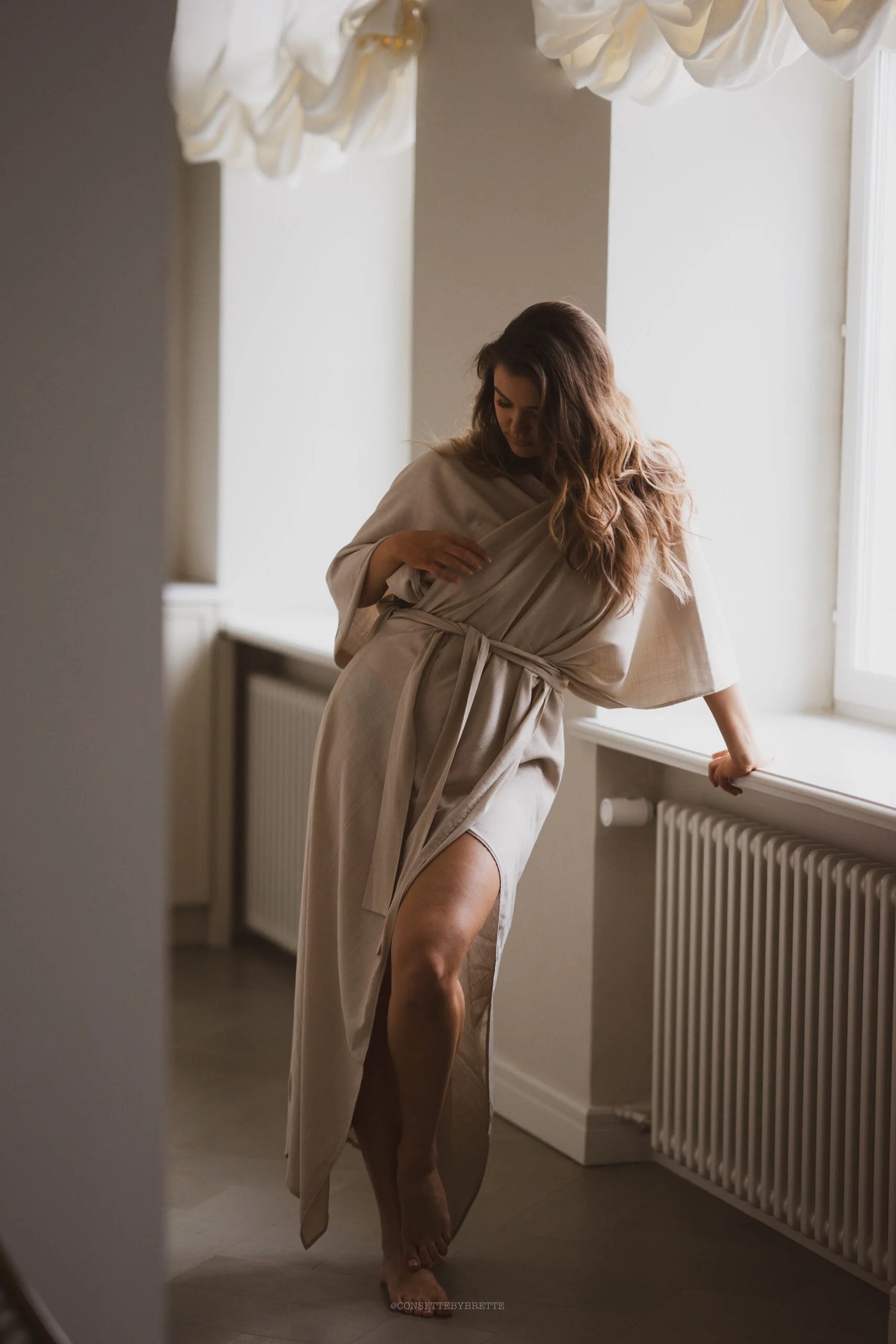 Woman in a beige robe standing by a window in a softly lit room.