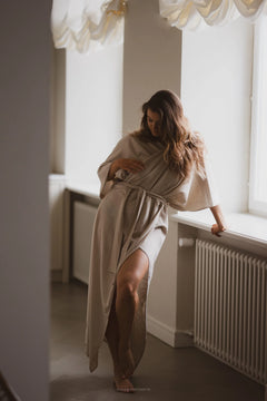Woman in a beige robe standing by a window in a softly lit room.