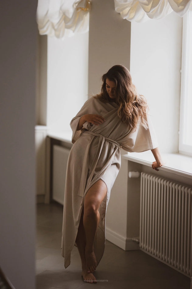 Woman in a beige robe standing by a window in a softly lit room.