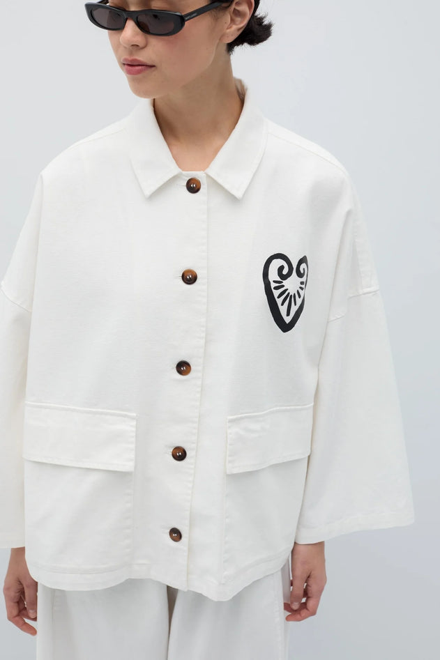 Person wearing a white jacket with a black heart design and brown buttons on a light gray background