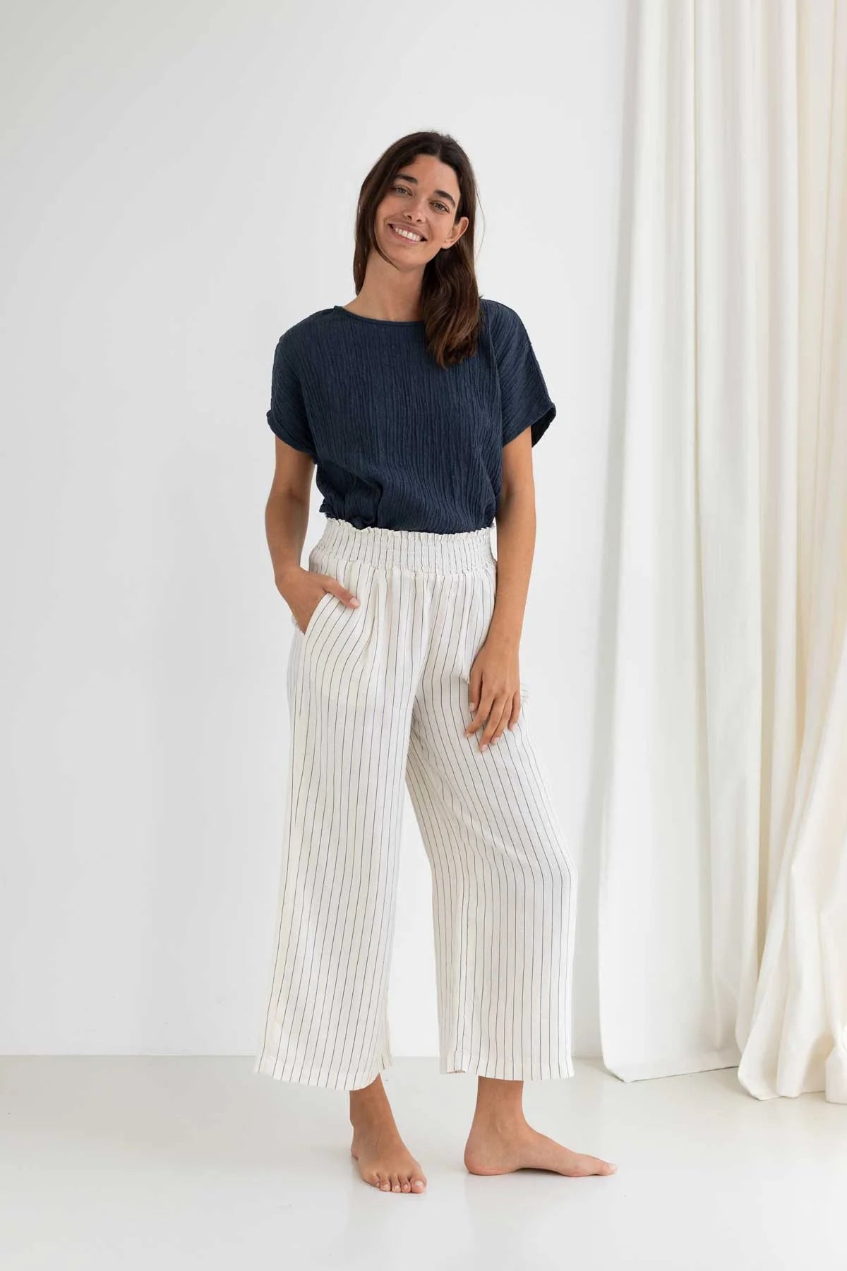Woman wearing a navy top and white striped pants standing against a white curtain.