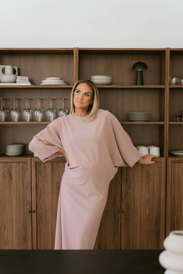 Woman in a pink dress standing in front of a wooden shelf with various items.