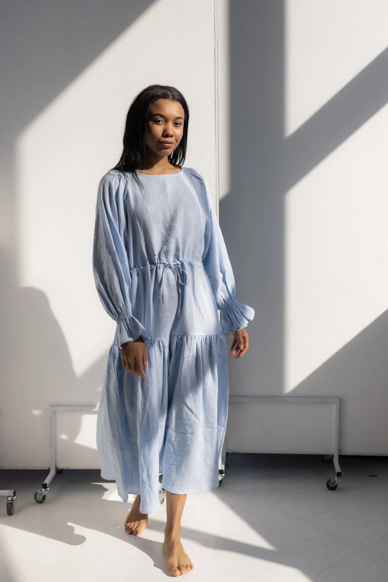 Woman wearing a light blue dress standing in a room with white walls and shadows.