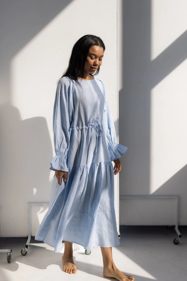 Woman wearing a light blue dress standing in a room with sunlight casting shadows on the wall.