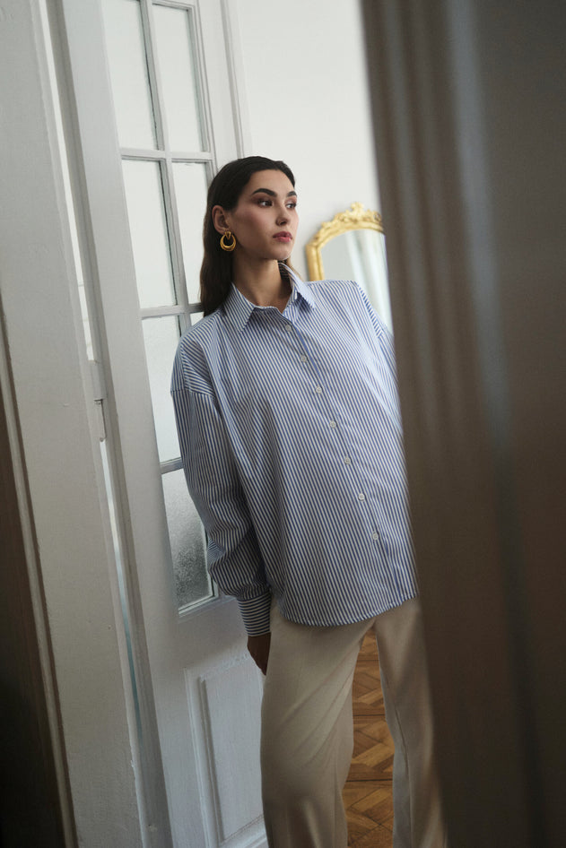 Woman wearing a blue checkered shirt and beige pants standing in a room with a white door and gold-framed mirror.
