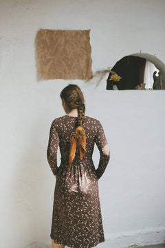 Person wearing a brown dress with white patterns, standing in front of a white wall.