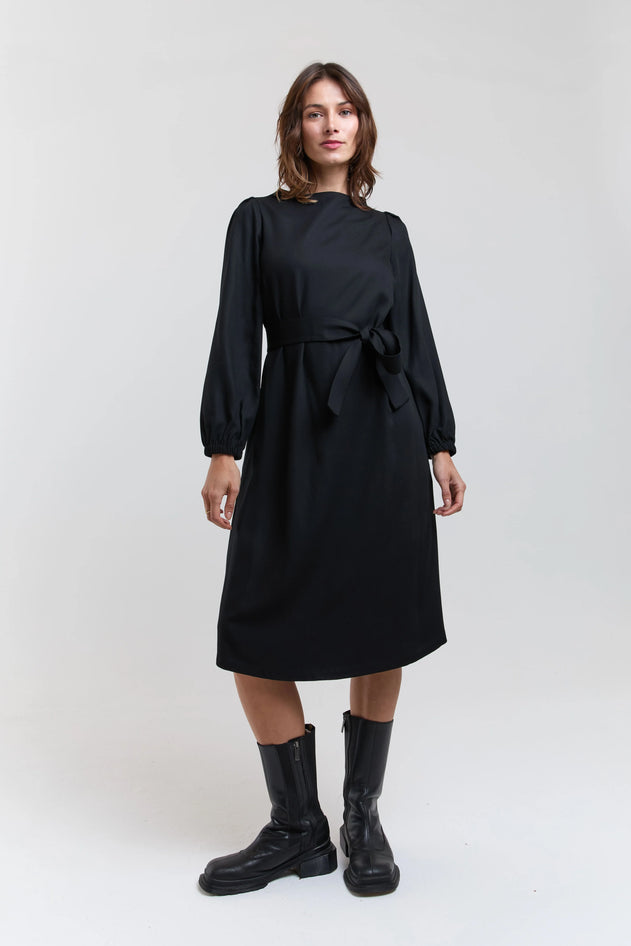 Woman wearing a black dress with long sleeves and a belt, standing against a white background.