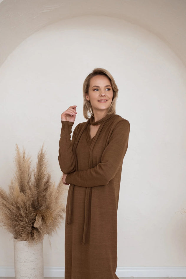 Woman wearing a brown dress standing against a plain wall with decorative elements.