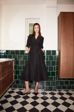 Woman in a black dress standing in a bathroom with green tiled walls and a checkered floor.