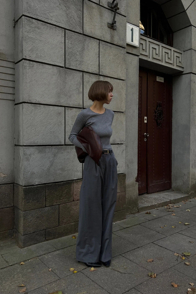 Person standing against a stone building wall holding a brown leather jacket.