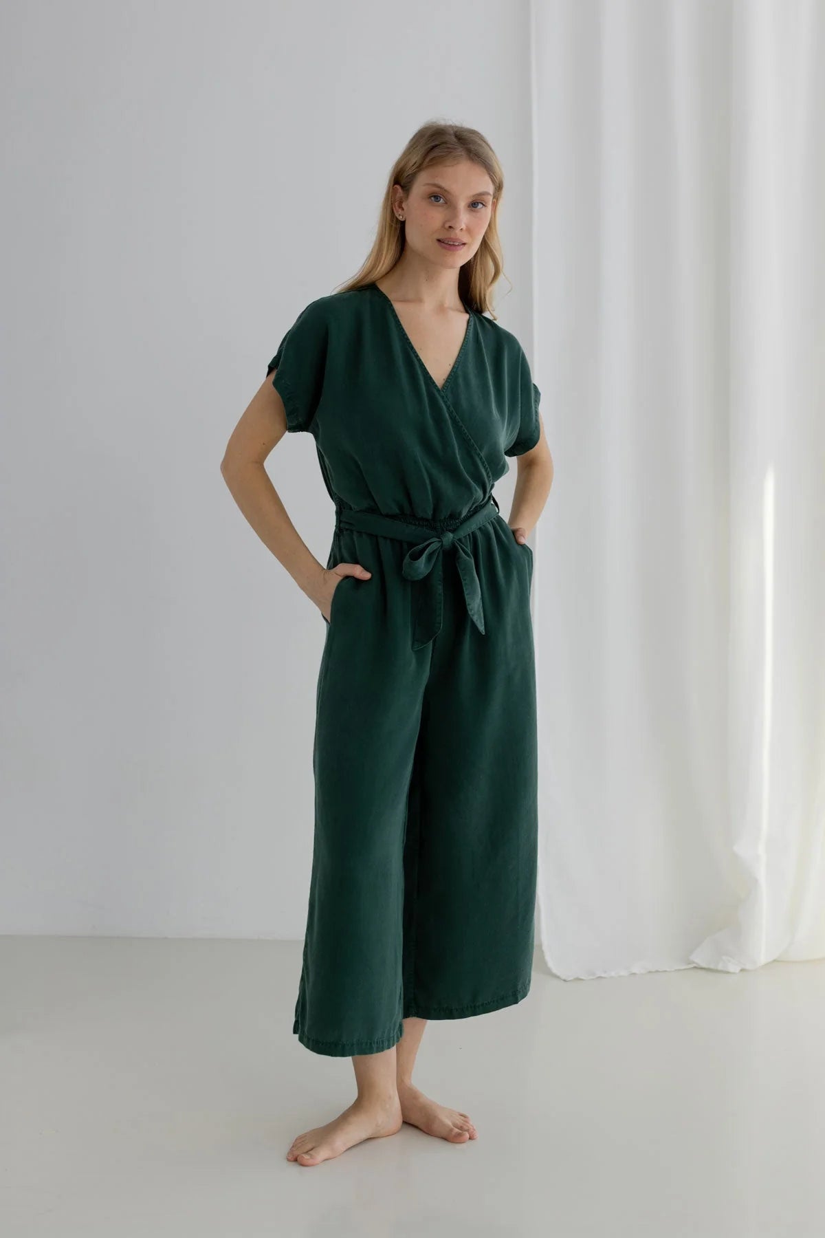 Woman wearing a green jumpsuit standing against a white curtain.