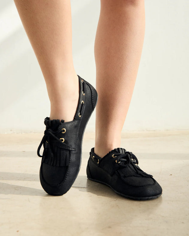 Black loafers with fringe details worn by a person on a light wooden floor.