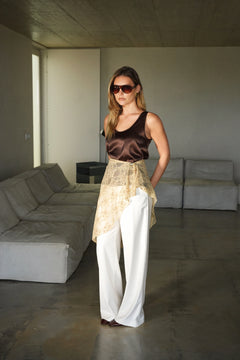 Woman standing in a modern living room wearing a brown top and white pants.