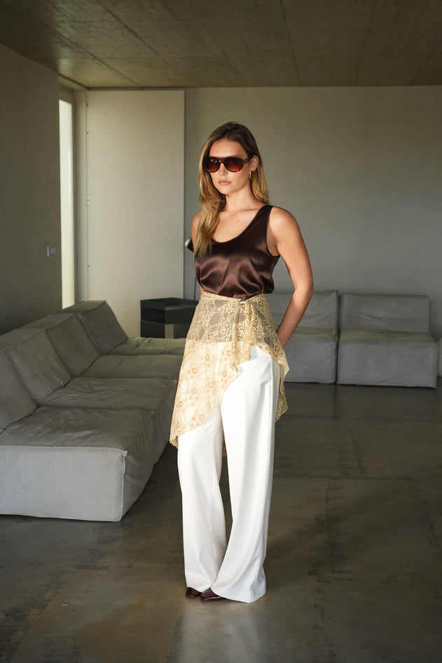 Woman standing in a modern living room wearing a brown top and white pants.