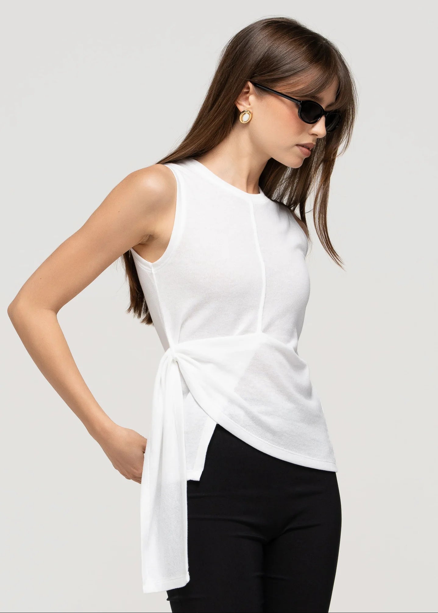 Woman wearing a white sleeveless top with a knot detail and black pants on a light gray background