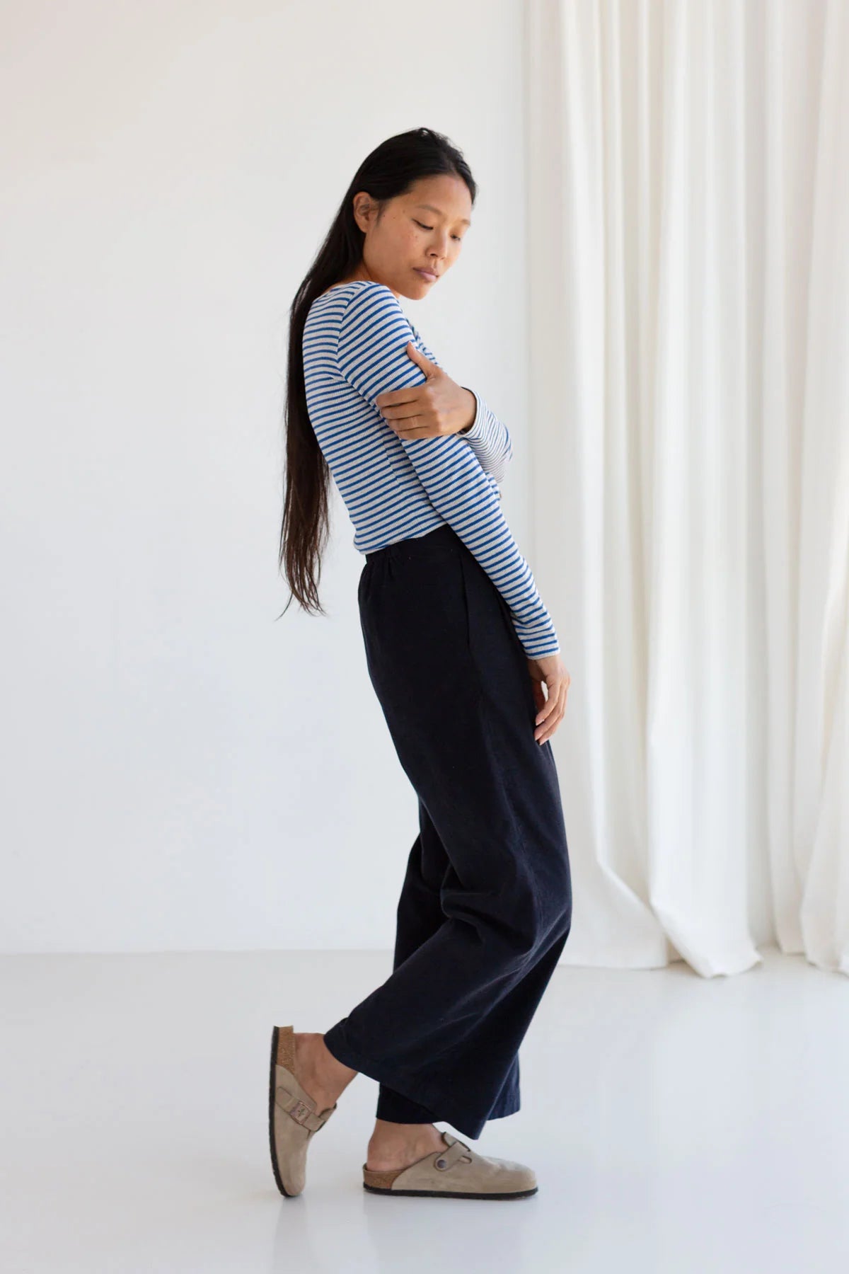 Woman wearing a striped shirt and dark pants standing against a white curtain.