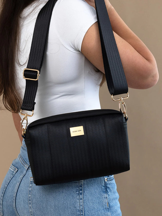 A black textured shoulder bag with a gold-tone logo plaque and adjustable strap, worn crossbody by a model in a white top and blue jeans against a beige background.