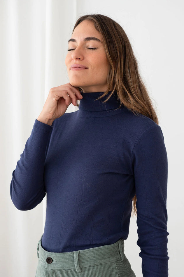 A woman wearing a navy blue ribbed mock neck turtleneck sweater, paired with olive green corduroy pants, posing against a white curtain background.