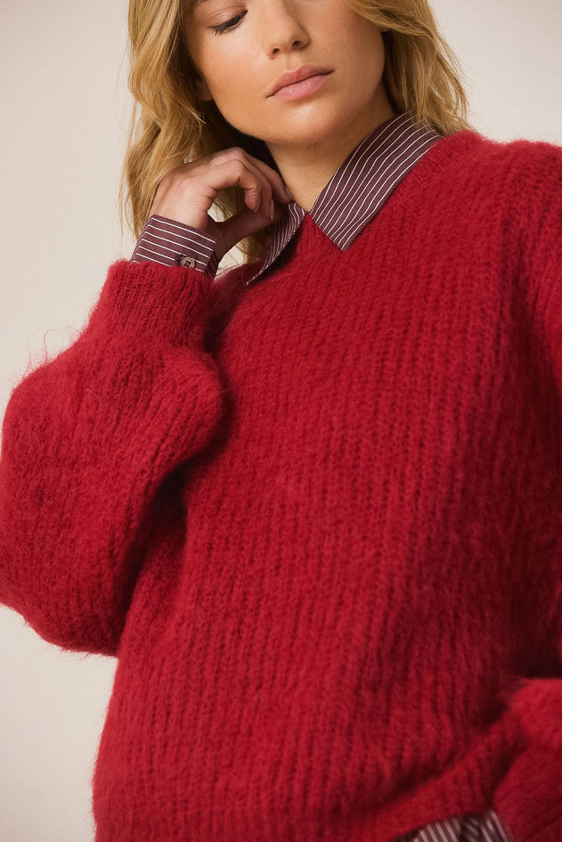 A close-up of a woman wearing a chunky, fuzzy red knit sweater layered over a collared shirt with a burgundy and beige stripe pattern.