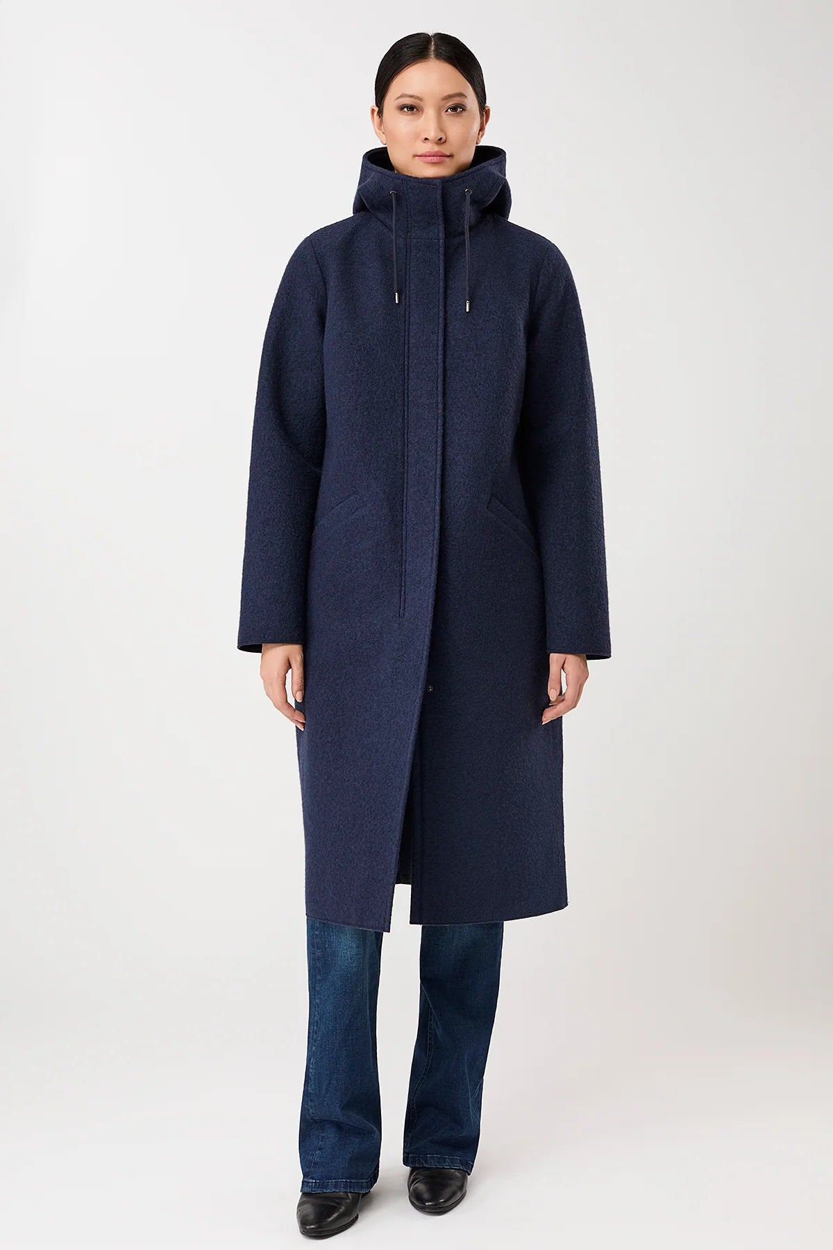 A model wearing a long navy blue hooded coat with a high collar, paired with blue jeans and black shoes, standing against a light gray studio background.
