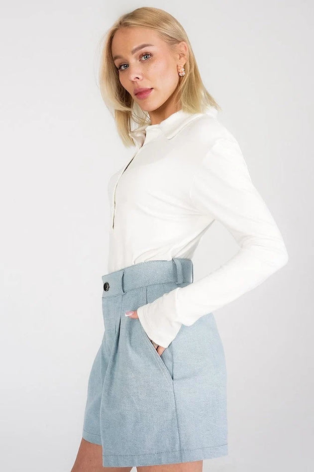 A blonde model wearing a white long-sleeve collared top and a light blue high-waisted pleated mini skirt with a matching belt, posed against a light gray studio background.