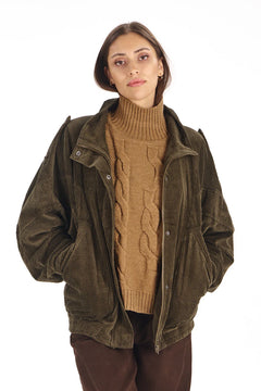 A woman wearing an olive green corduroy jacket over a tan cable-knit turtleneck sweater and dark brown pants, photographed against a plain white background.