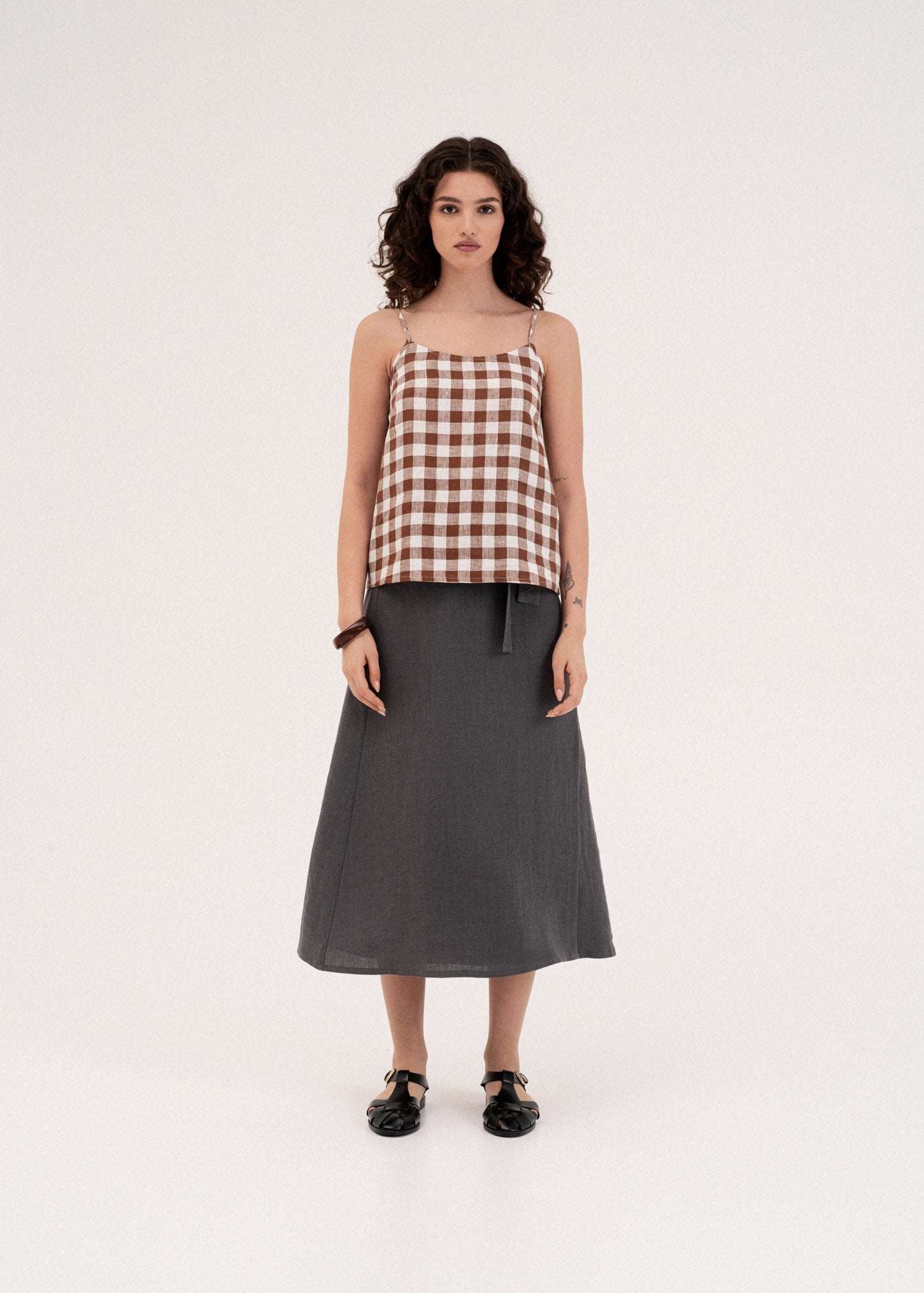 A model stands against a plain white studio background wearing a brown and white gingham patterned camisole top with thin straps and a dark gray midi skirt.