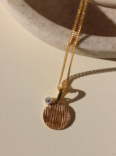Gold necklace with a textured pendant and diamond on a beige surface
