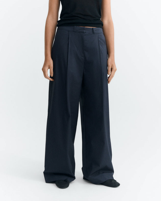 A model wearing a pair of dark navy wide-leg pleated trousers, paired with a black top and black shoes, photographed against a clean white studio background.