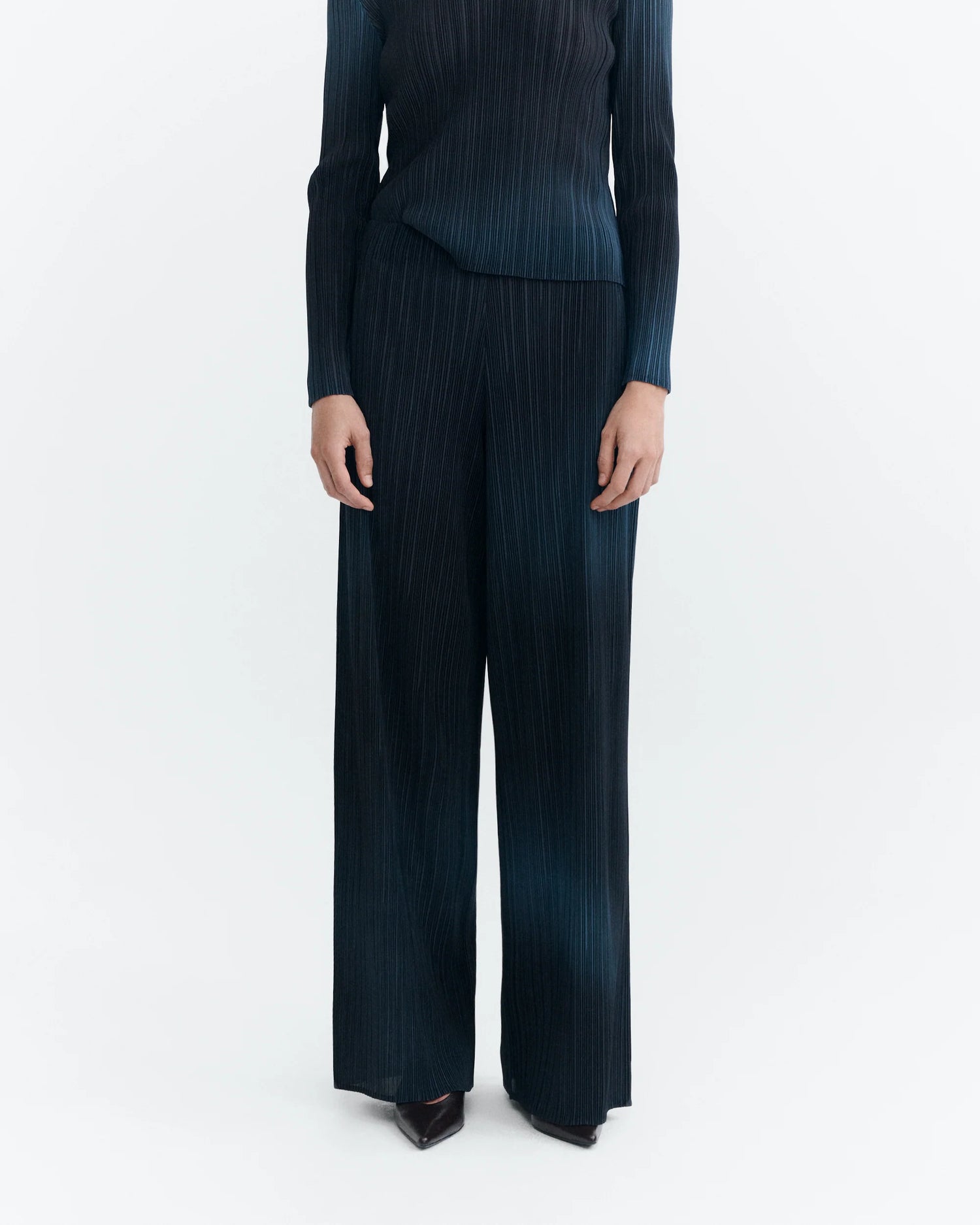 A model wearing a dark blue pleated two-piece set consisting of a long-sleeve top and matching wide-leg trousers, shown from the neck down on a light gray background.