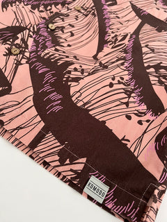 A close-up of the SPINDRIFT - Organic Cotton Shirt - Pink Wave, showing its abstract dark brown and purple wave print, button details, and a small KOMODO fabric tag on the hem.