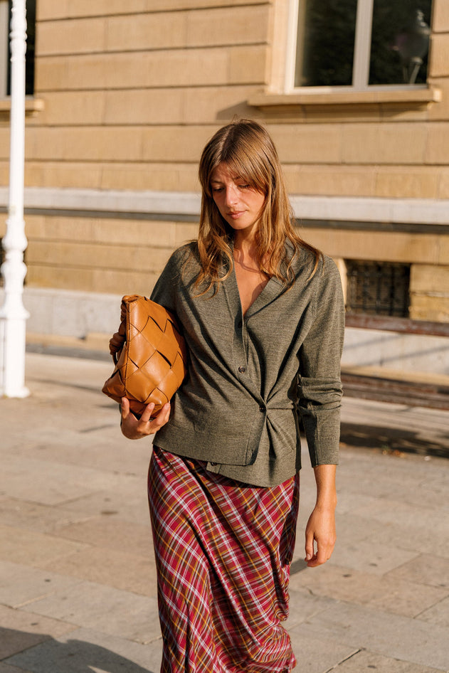 A woman walking outdoors on a sunny day, wearing a dark olive green button-up shirt and a red and green plaid skirt, while holding a brown woven leather clutch bag.